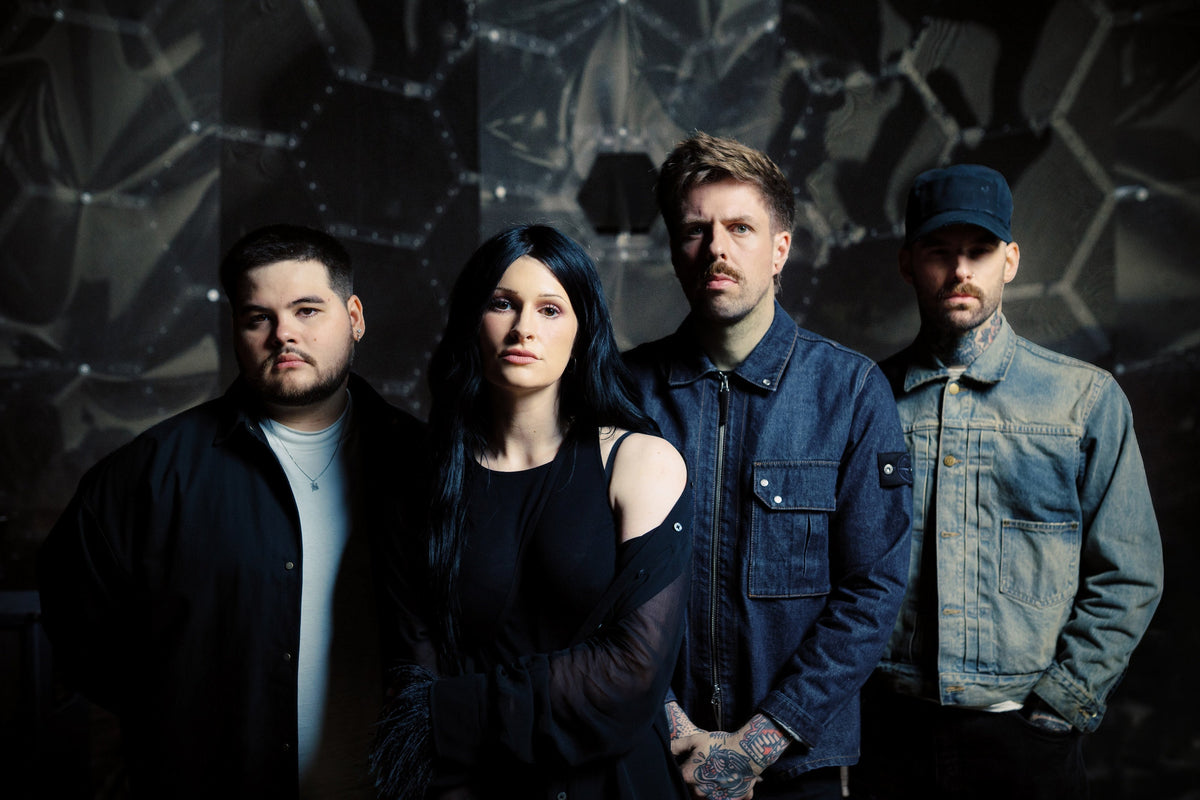 Four people stand in front of a corrugated metal wall with graffiti, looking directly at the camera. The image is black and white, giving it a serious and moody atmosphere.