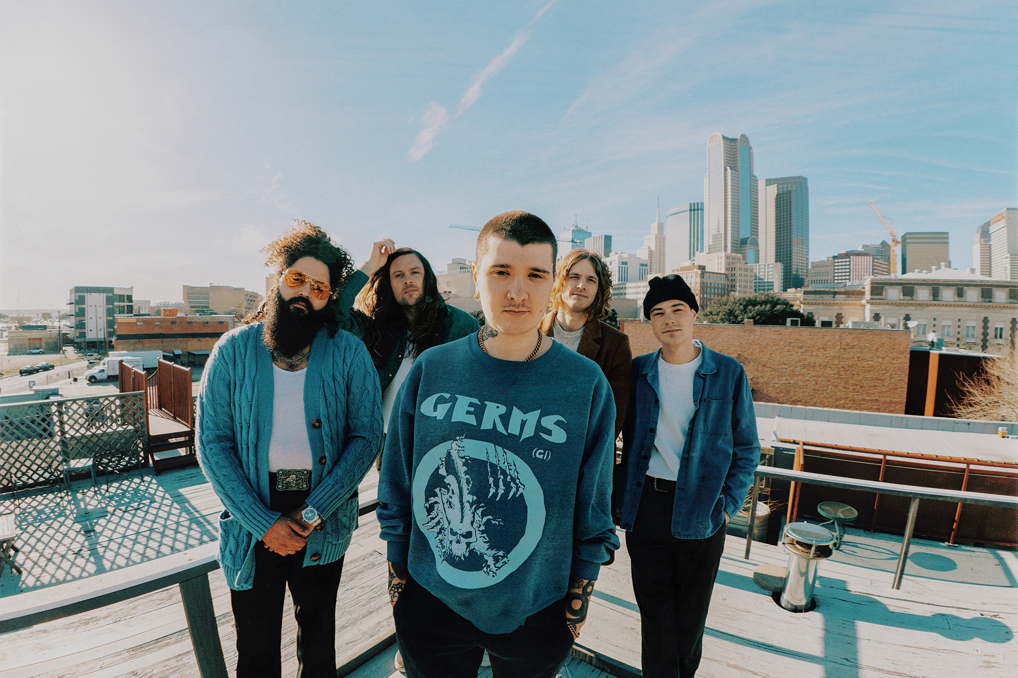 Five people stand on a rooftop with a city skyline in the background. The person in front wears a GERMS sweatshirt; others wear casual clothing. Its a sunny day with clear skies.