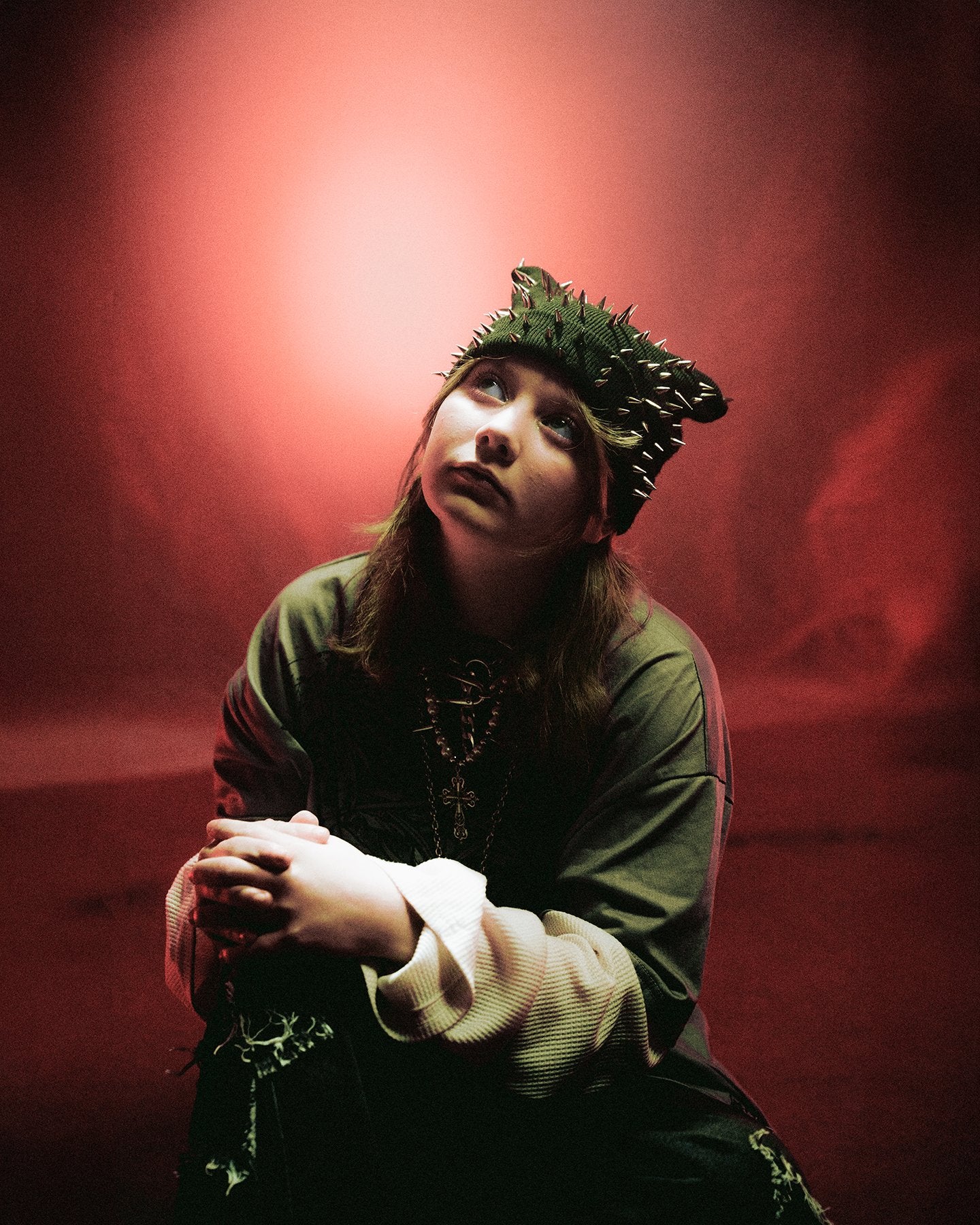 A person wearing a spiked headpiece and layered clothing sits with hands clasped, looking upward. The background is dark with a dramatic red light glowing behind them.