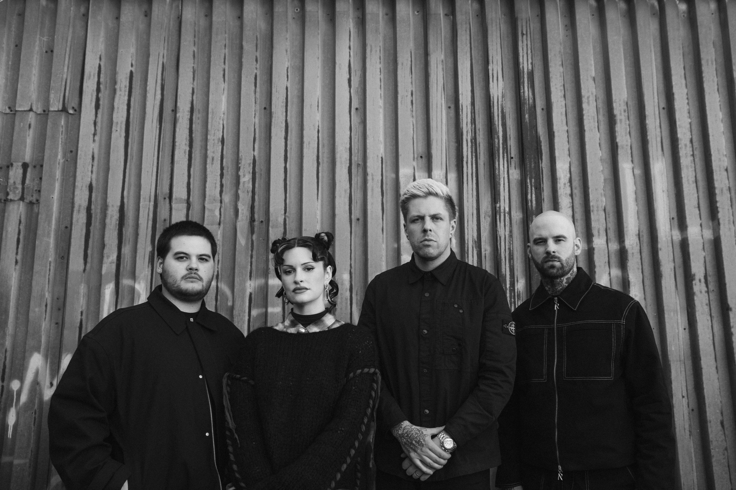 Four people stand in front of a corrugated metal wall with graffiti, looking directly at the camera. The image is black and white, giving it a serious and moody atmosphere.