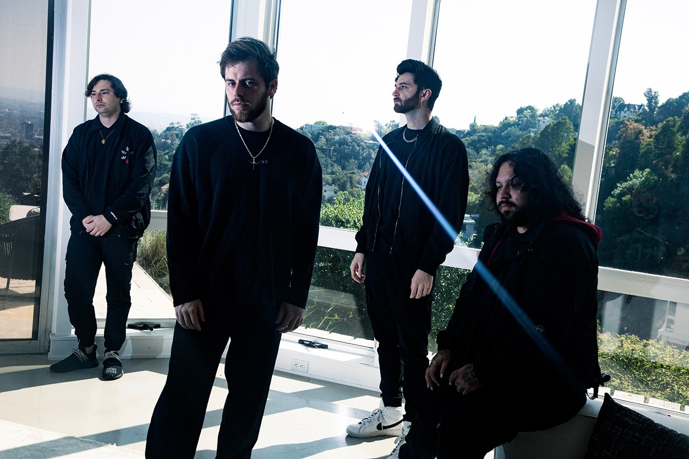Four men dressed in dark clothing stand in a bright room with large windows overlooking greenery; a lens flare crosses the image diagonally.