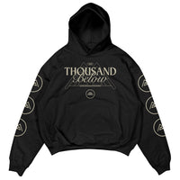 Black Thousand Below hoodie featuring premium Shaka Wear material, a geometric mountain and band logo on the front, plus circular mountain logos running down each sleeve—an eye-catching printed garment.