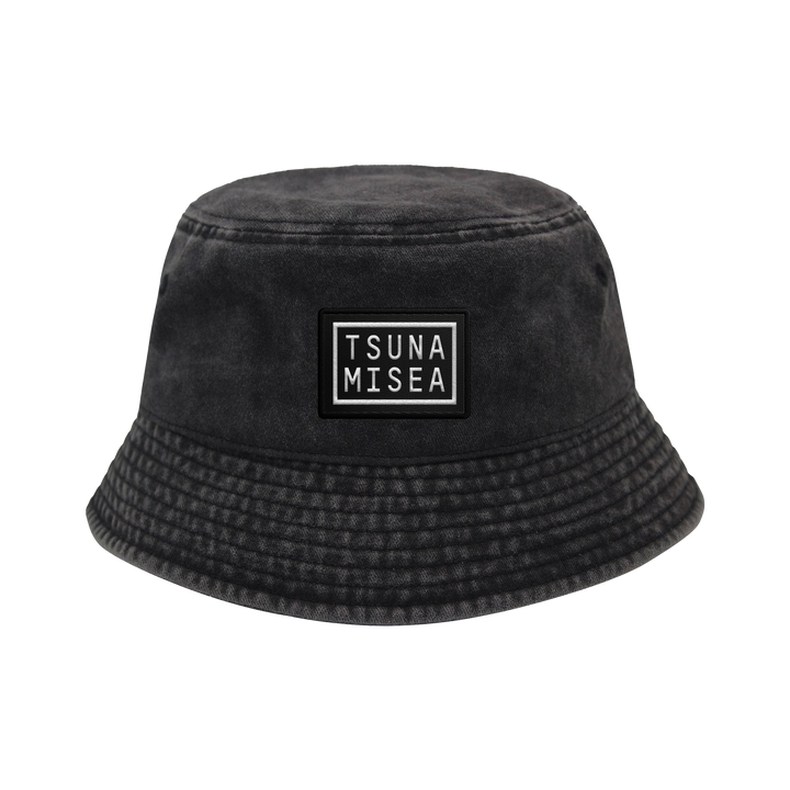 A Spiritbox Pigment Black Bucket Hat with a rectangular "TSUNA MISEA" patch on the front in white uppercase letters, featuring slightly textured fabric and a downward-sloping brim.
