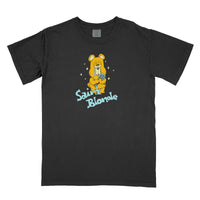 This black Saint Blonde t-shirt features a comfort colors design with a printed sad yellow bear holding a blue ball, sparkles, and “Saint Blonde” in light blue stylized letters below the illustration.