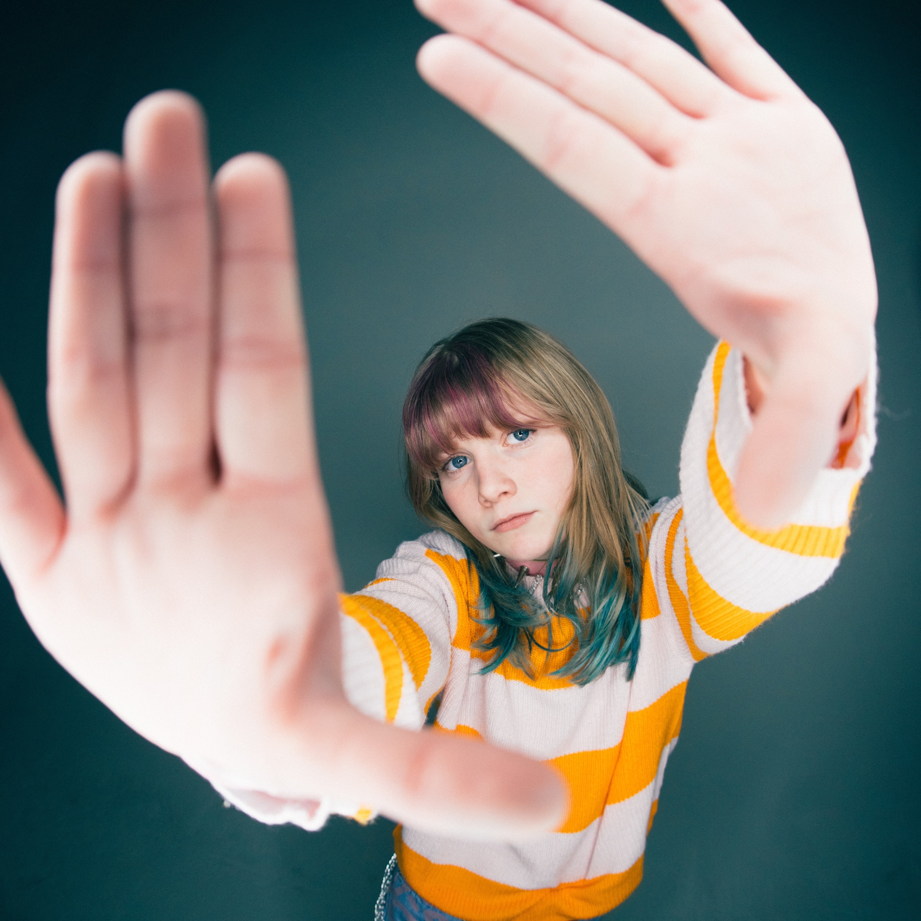 A young person with long hair and a white-and-yellow striped sweater holds their hands up toward the camera, framing their face with their fingers, against a plain dark background.