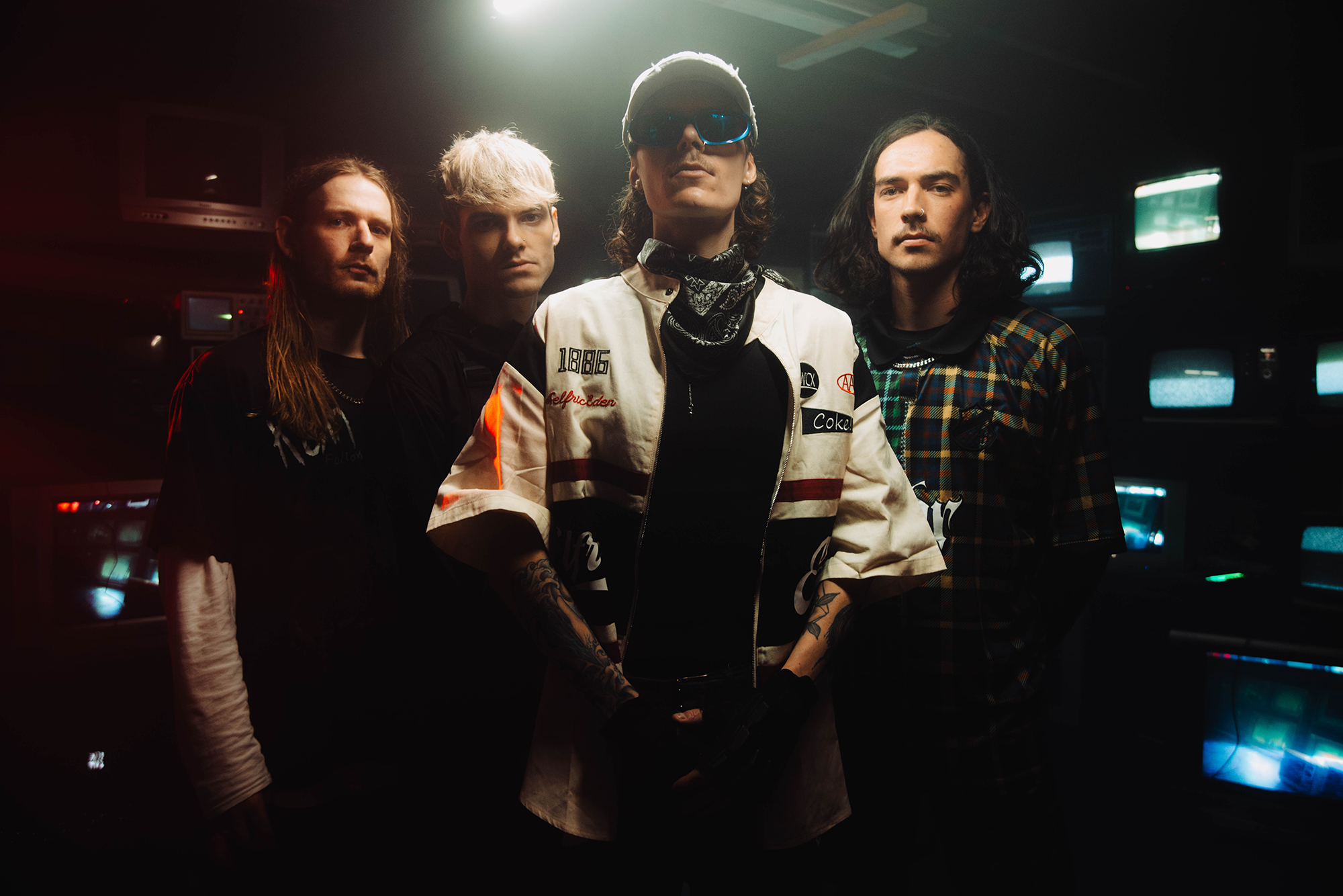 Four young adults stand together in a dimly lit room with glowing old televisions in the background. The person in front wears sunglasses, a white jacket, and a cap, while the others wear casual dark clothing.