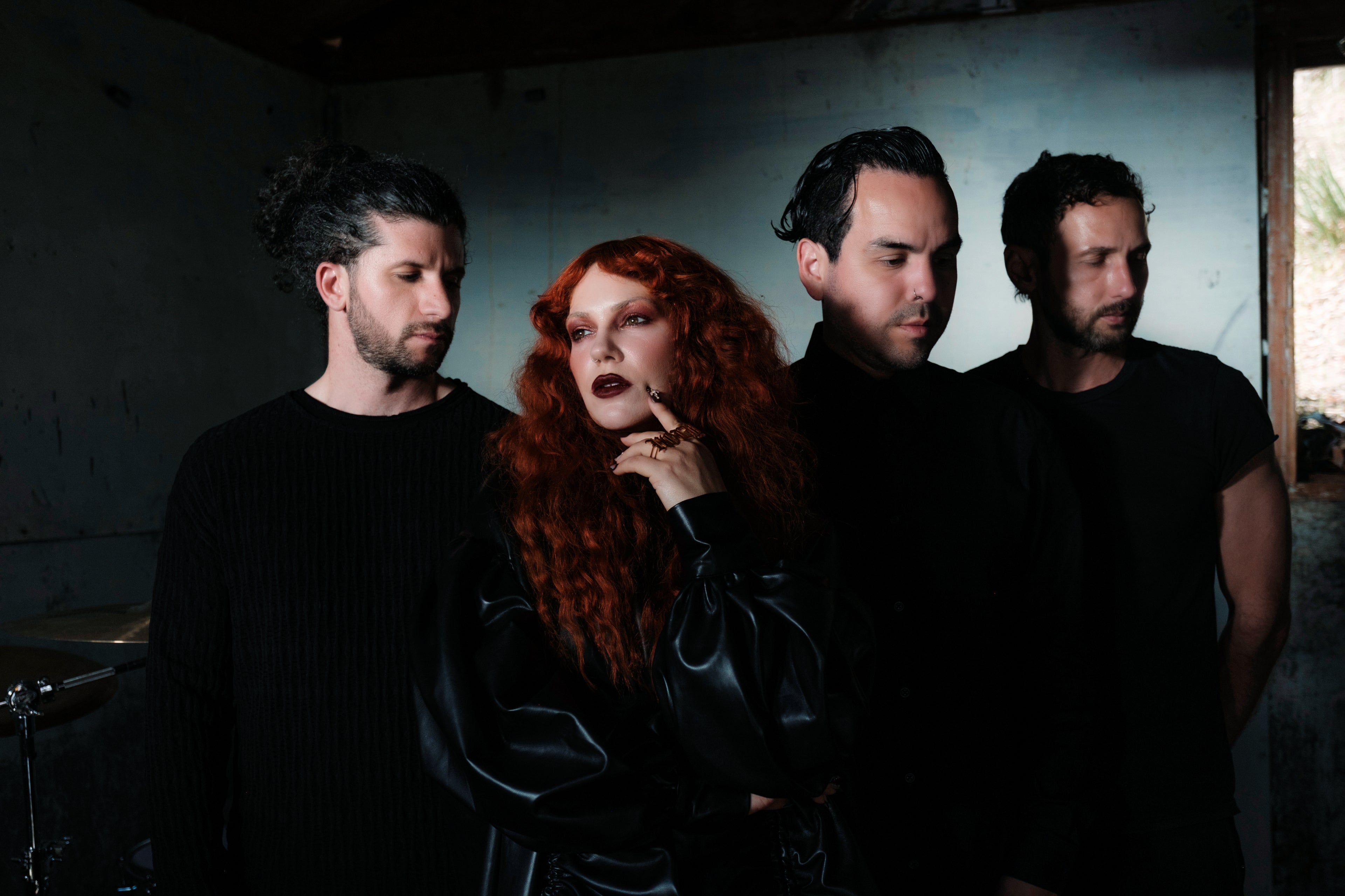 Four people stand together in a dimly lit room; three men wear black and look away, while a woman with long red curly hair and dark lipstick stands in front, gazing forward with her hand on her chin.