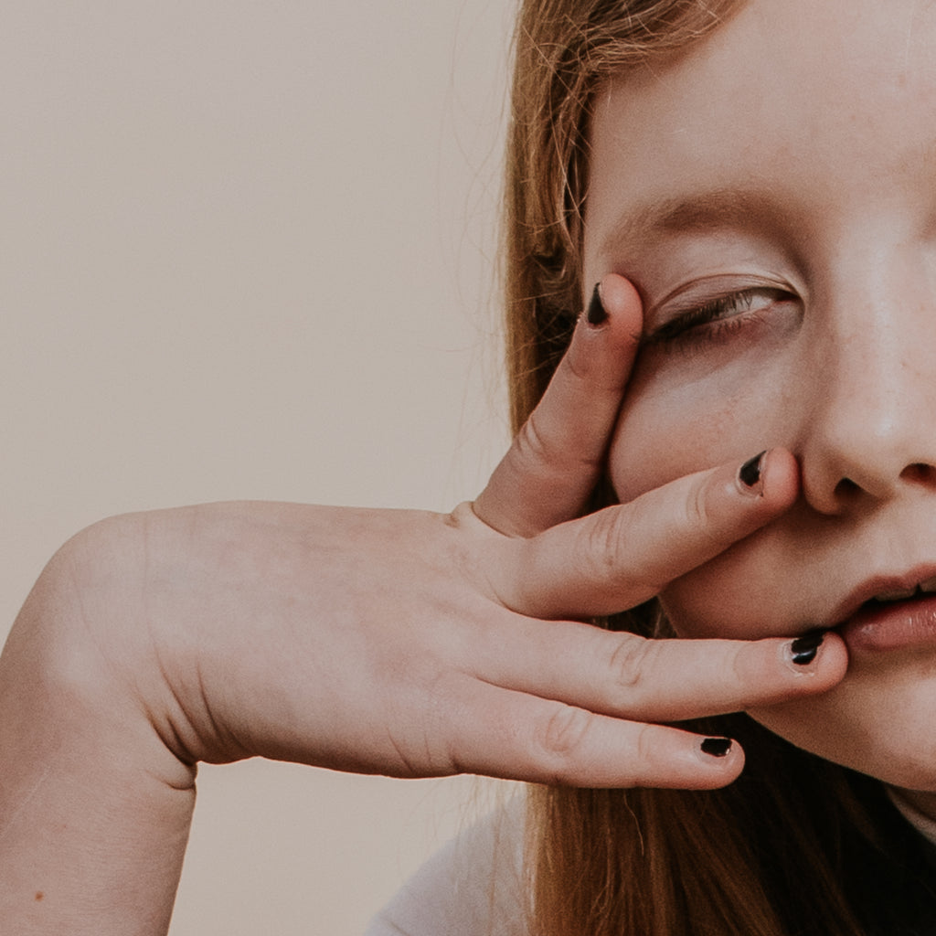 A close-up of a child with light skin and long hair, holding three fingers with chipped black nail polish to their face, partially covering their eye and looking upward. The background is plain and neutral.