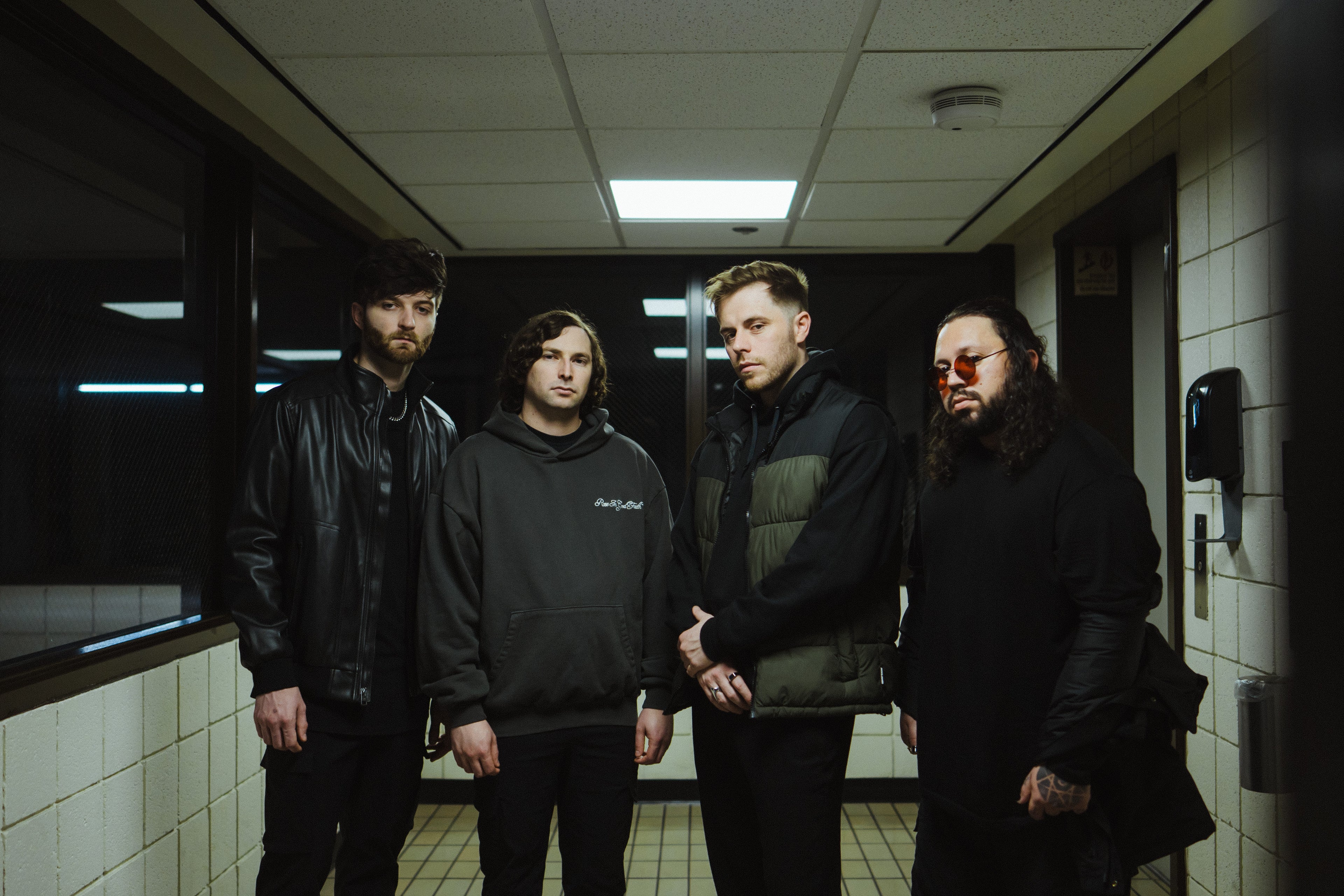 Four men stand in a tiled hallway with fluorescent lighting; they wear dark, casual clothing and have serious expressions. The background features windows, doors, and a soap dispenser on the wall.
