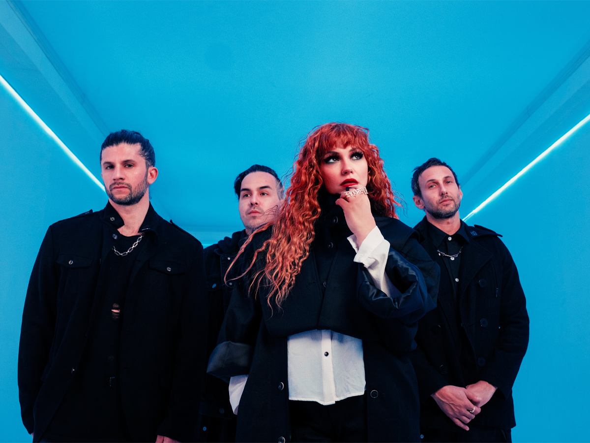Four people stand in a blue-lit room. The woman in front has curly red hair and wears black and white clothing, while three men in black outfits stand behind her, all looking forward with serious expressions.