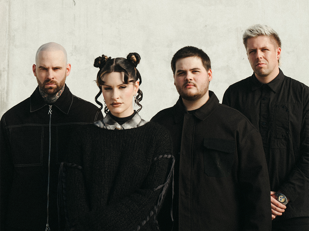 Four people stand side by side against a plain, light-colored background, all dressed in dark clothing. The person on the left is bald, and the person second from left has space buns and curled bangs.